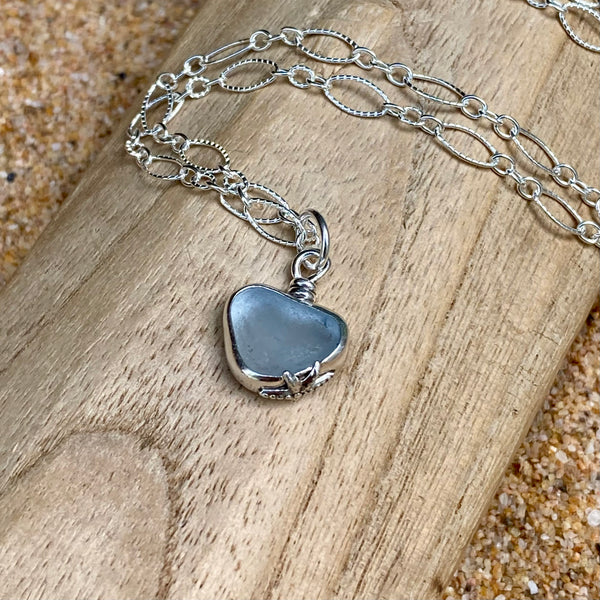 A sterling silver chain necklace with an ice  heart-shaped sea glass pendant, displayed on a piece of natural wood with sand in the background.