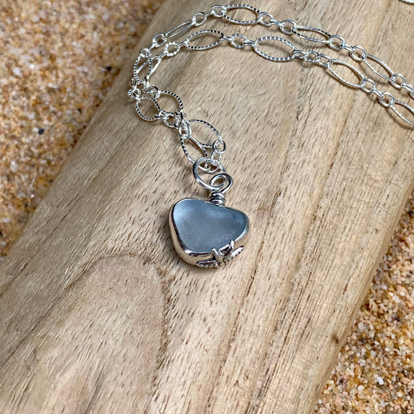 A sterling silver chain necklace with an ice  heart-shaped sea glass pendant, displayed on a piece of natural wood with sand in the background.