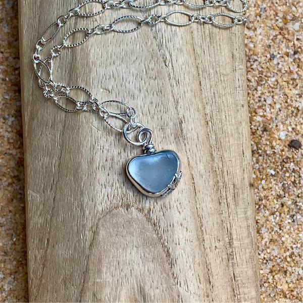 A sterling silver chain necklace with an ice  heart-shaped sea glass pendant, displayed on a piece of natural wood with sand in the background.