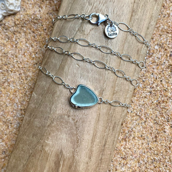 A sterling silver chain necklace with a light teal grey heart-shaped sea glass pendant, displayed on a wooden slice over a sandy background.