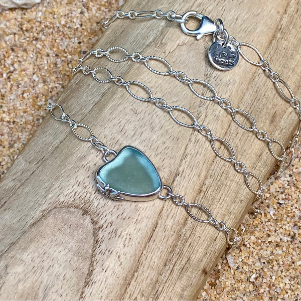A sterling silver chain necklace with a light teal grey heart-shaped sea glass pendant, displayed on a wooden slice over a sandy background.