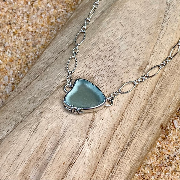 A sterling silver chain necklace with a light teal grey heart-shaped sea glass pendant, displayed on a wooden slice over a sandy background.