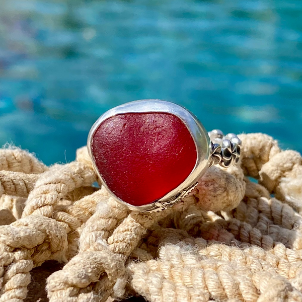 A silver ring with a large, polished red sea glass cabochon, resting on a bundle of beige rope with a blurred blue ocean background.