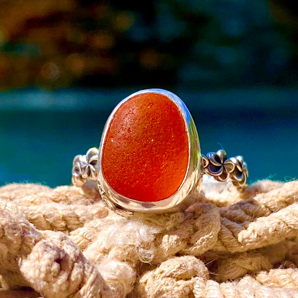 A silver ring with a large, oval, translucent orange-red sea glass cabochon. displayed on a textured beige rope with a blurred blue-green background.