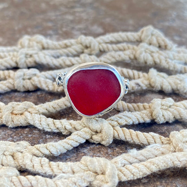 A silver ring with a large, rounded red sea glass, displayed on a knotted beige rope surface.