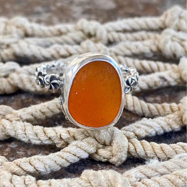 A silver ring with a large, rounded orange sea glass cabochon, displayed on a bed of thick beige rope.