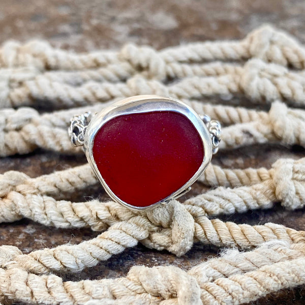 A sterling silver ring with a large, irregularly shaped deep red sea glass cabochon, displayed on a rustic background of cream-colored rope.